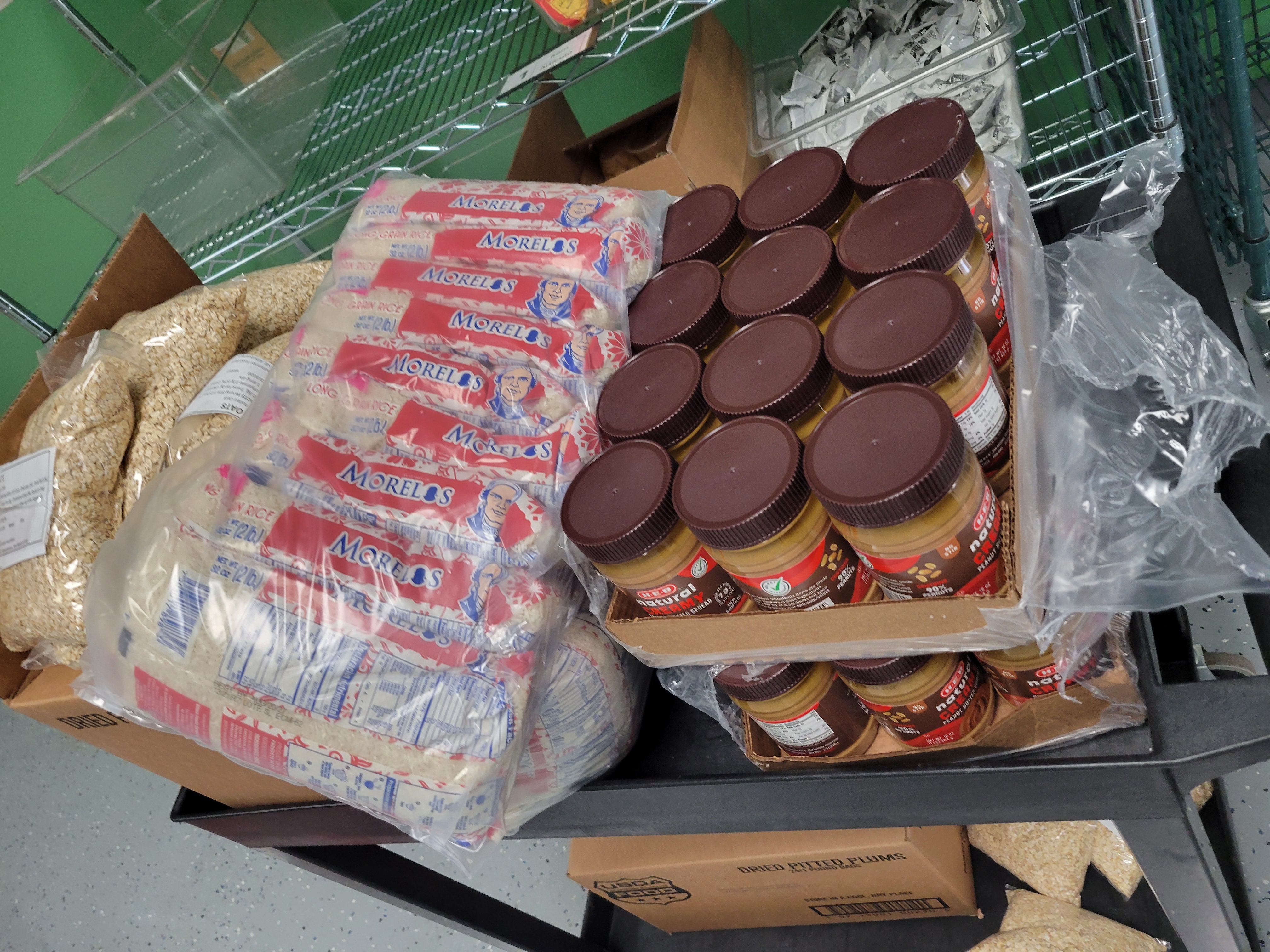 Boxes of bags of oatmeal, bags of rice, and trays of peanut butter on top of the shelf of a shelf cart with the bottom filled with a box of dried pitted plums and bags of oats.